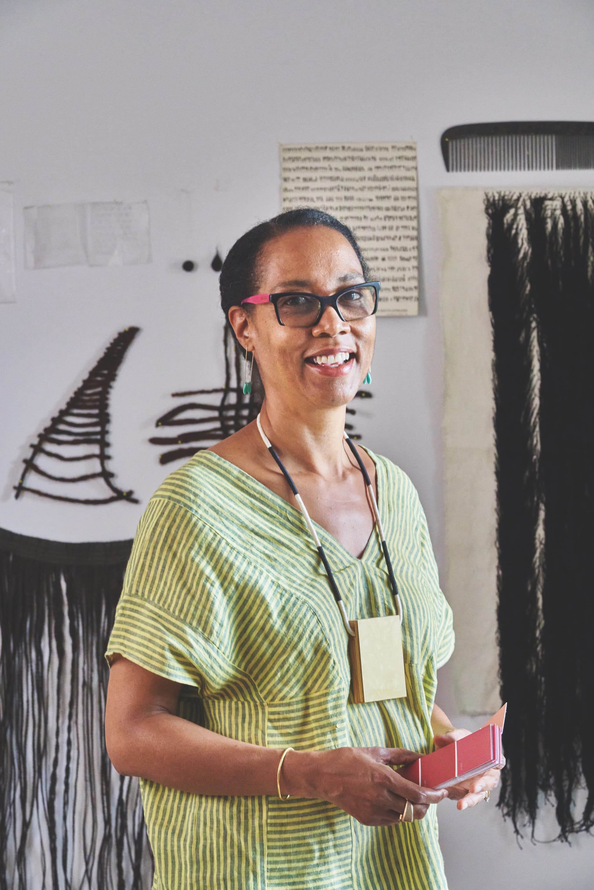 A portrait of Sonya Clark, wearing green, in her studio, standing in front of black and white artworks.