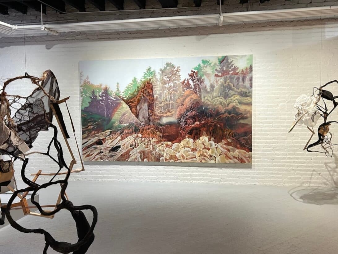 A landscape artwork and sculptures hung from the ceiling of a gallery space.